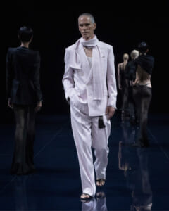 Male model in a pale pink/white suit and scarf walking a fashion runway against a dark backdrop.