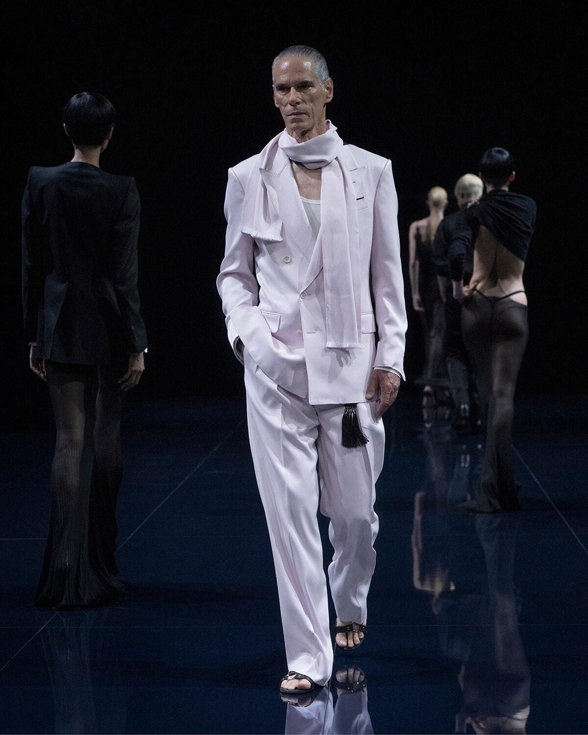 Male model in a pale pink/white suit and scarf walking a fashion runway against a dark backdrop.