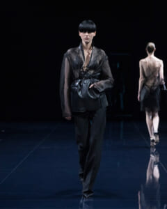 Model walking a dark runway in an all-black outfit: sheer lace top, wide belt, and a leather vest with a bag, with another model visible in the background.