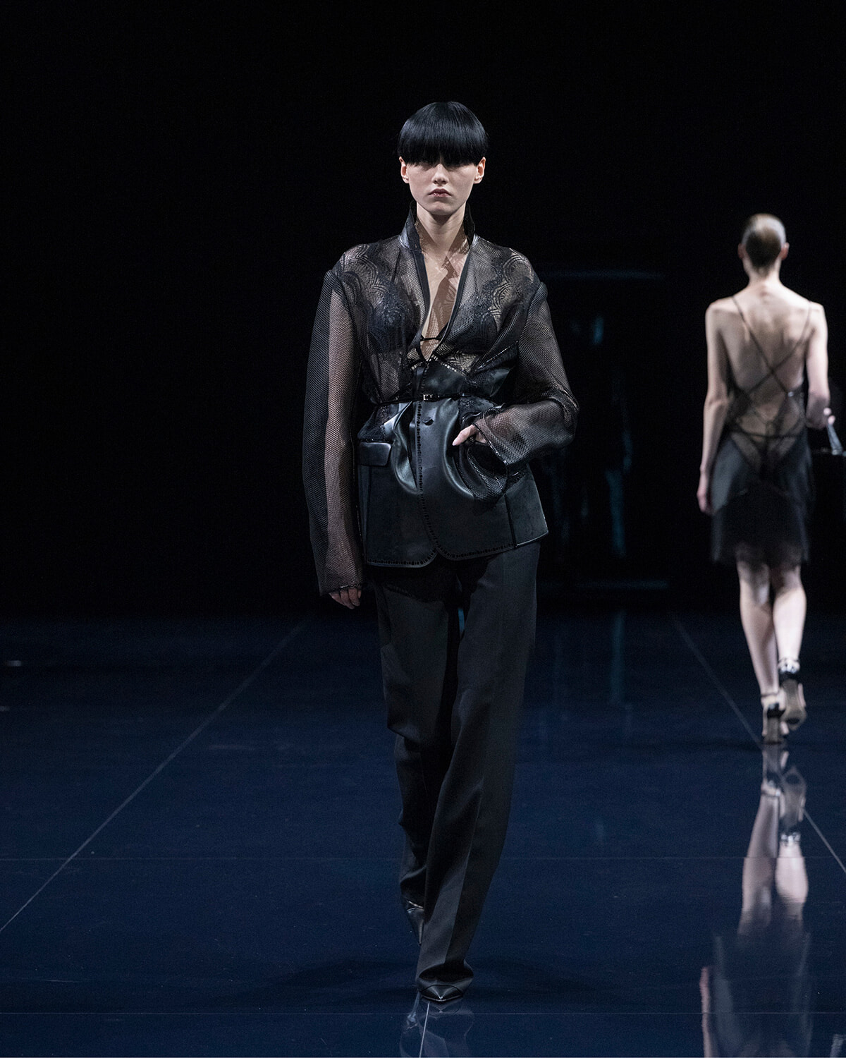 Model walking a dark runway in an all-black outfit: sheer lace top, wide belt, and a leather vest with a bag, with another model visible in the background.