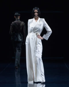 Female model in a white wrap gown with a belt stands on a dark runway, another model in black in the background.