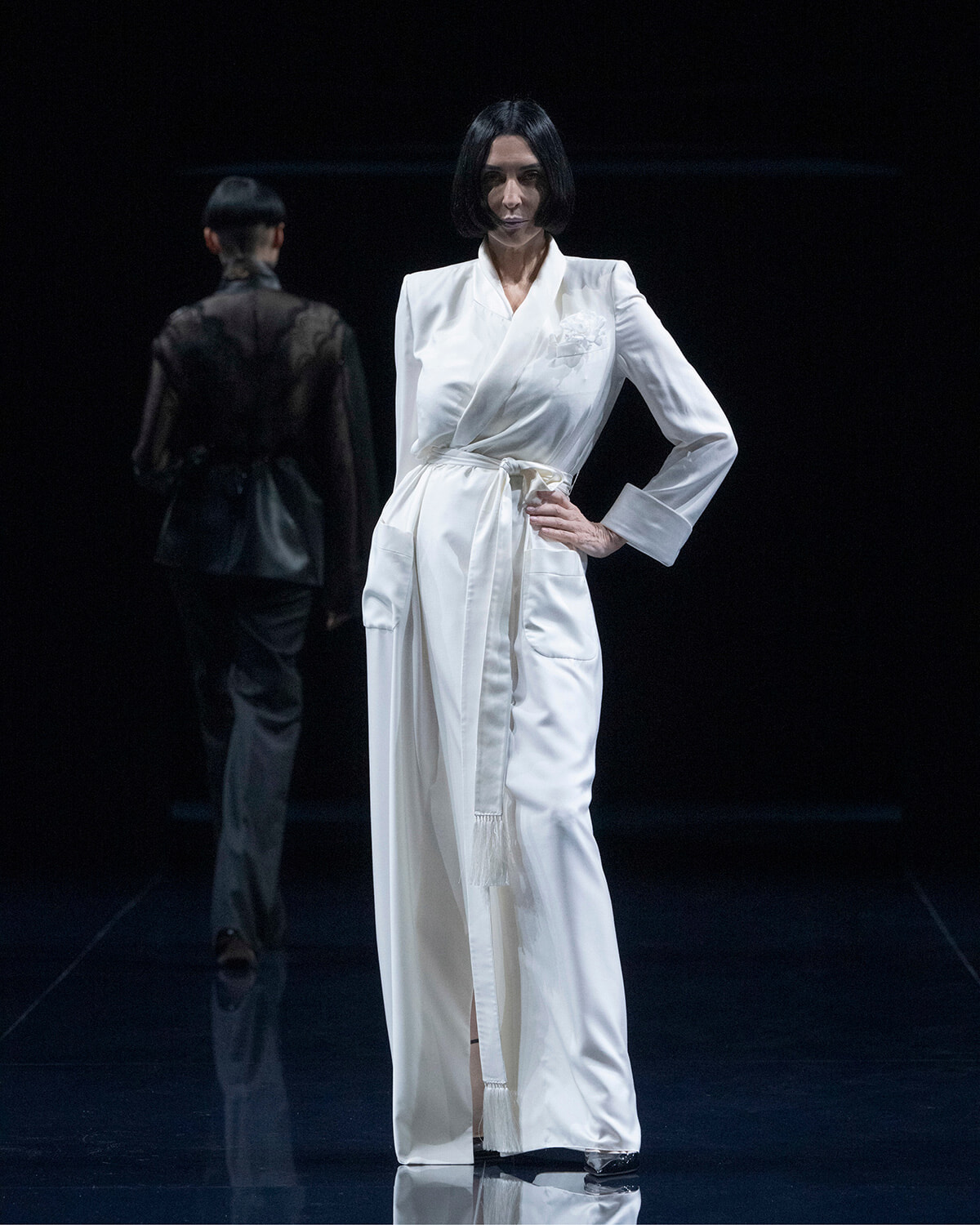Female model in a white wrap gown with a belt stands on a dark runway, another model in black in the background.