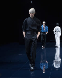 Model walks a dark runway wearing a black outfit; cropped, ice-blond bowl haircut and hands in pockets, glossy floor reflecting the scene