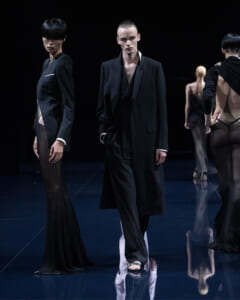 Group of fashion models on a dark runway wearing black, with a central male in a long coat and layered outfit; glossy floor reflections visible.