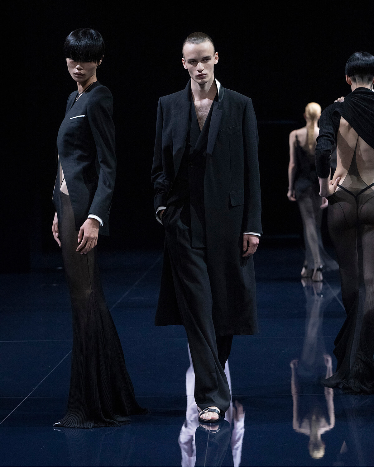 Group of fashion models on a dark runway wearing black, with a central male in a long coat and layered outfit; glossy floor reflections visible.