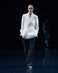 Male fashion model on a dark runway wearing a white blazer, black trousers, and black slide sandals, standing with hands in pockets.