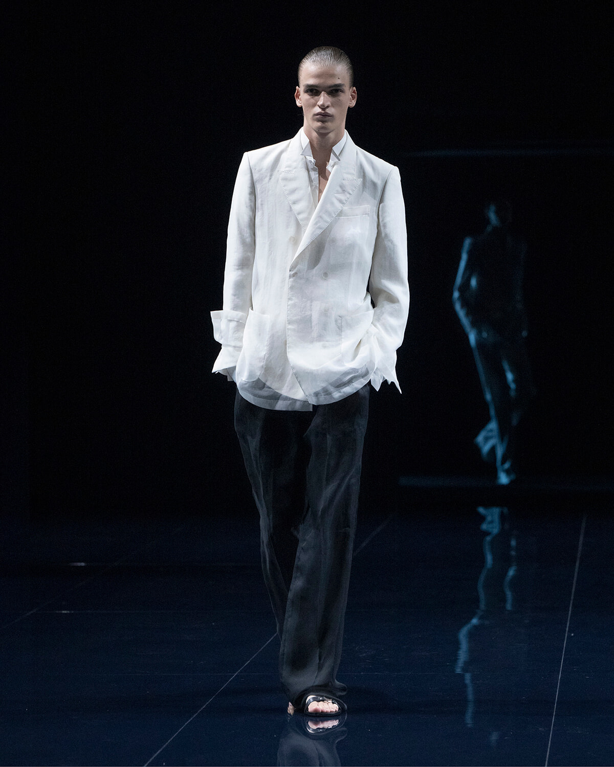 Male fashion model on a dark runway wearing a white blazer, black trousers, and black slide sandals, standing with hands in pockets.