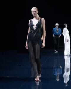 Model walks the runway in a sheer black lace jumpsuit with plunging neckline, dark backdrop behind her.