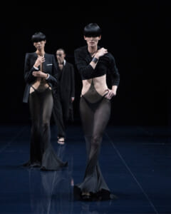 Models walk a dark runway in sheer black cut-out gowns with pleated, floor-length hems.