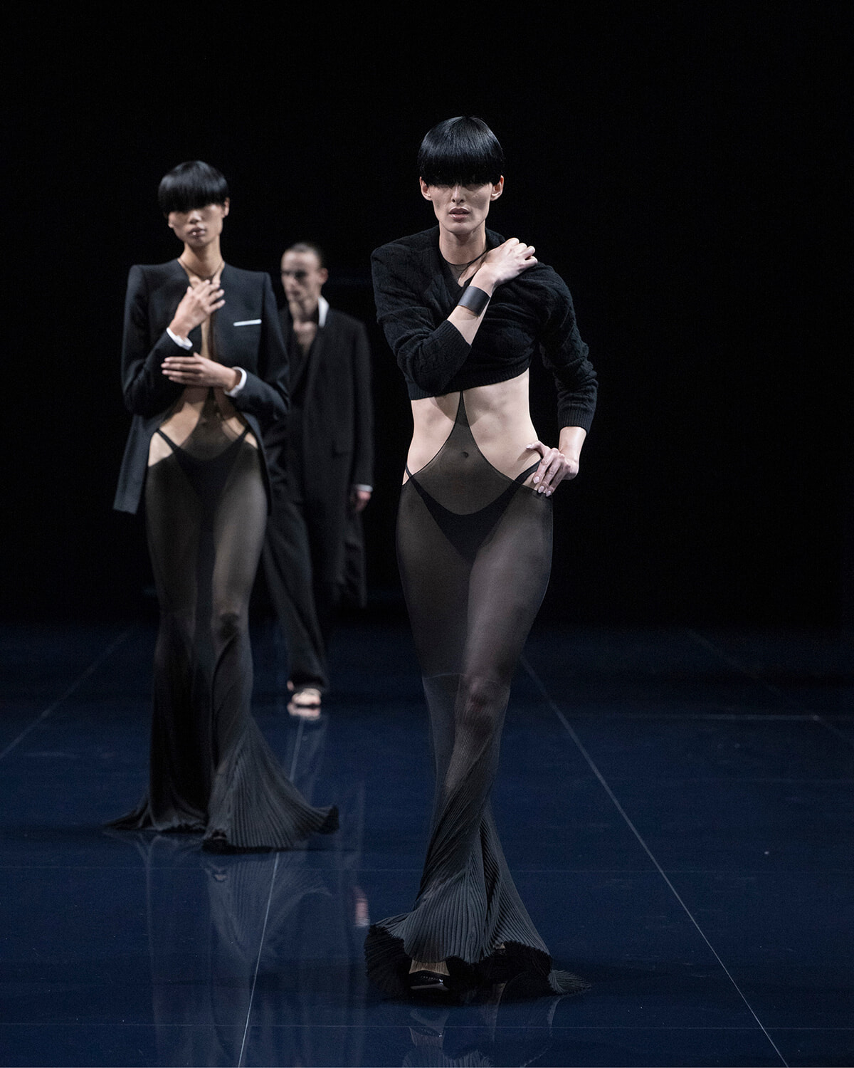 Models walk a dark runway in sheer black cut-out gowns with pleated, floor-length hems.