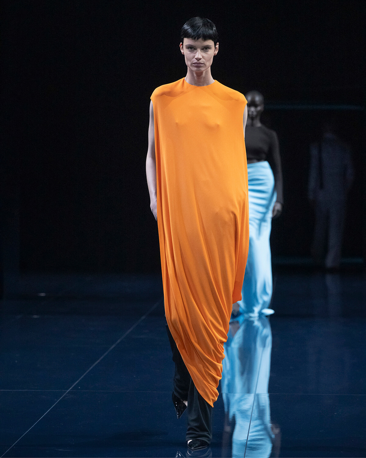 Model on a dark runway wearing a bright orange draped sleeveless dress, black pants and pointed shoes; second model in blue pants visible in the background.