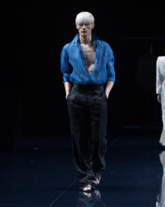 Model walking a runway in a bright blue sheer blouse worn open over a bare chest and loose black trousers, silver short hair.