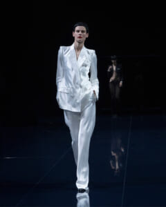 Model walking the runway in a glossy white pantsuit with a structured blazer on a dark catwalk.