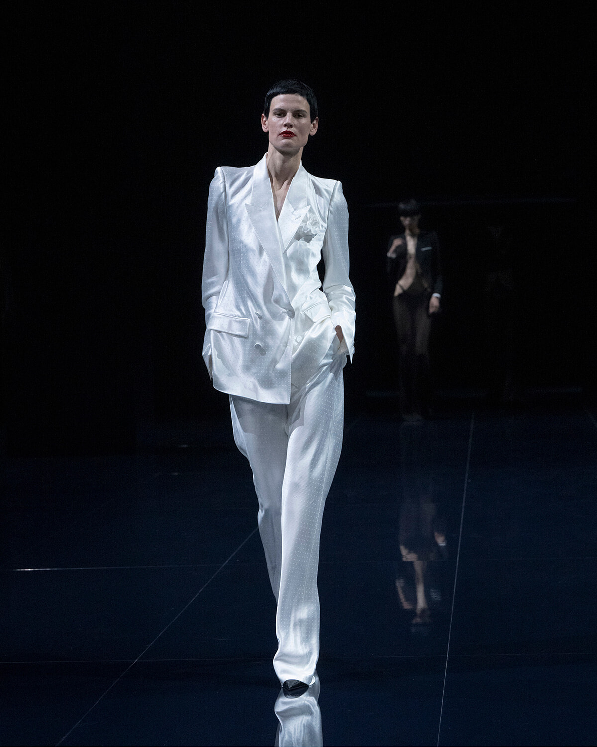 Model walking the runway in a glossy white pantsuit with a structured blazer on a dark catwalk.