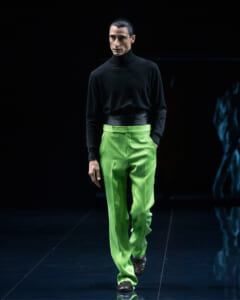 Male model walking the runway in a black turtleneck and bright lime-green trousers on a dark stage.