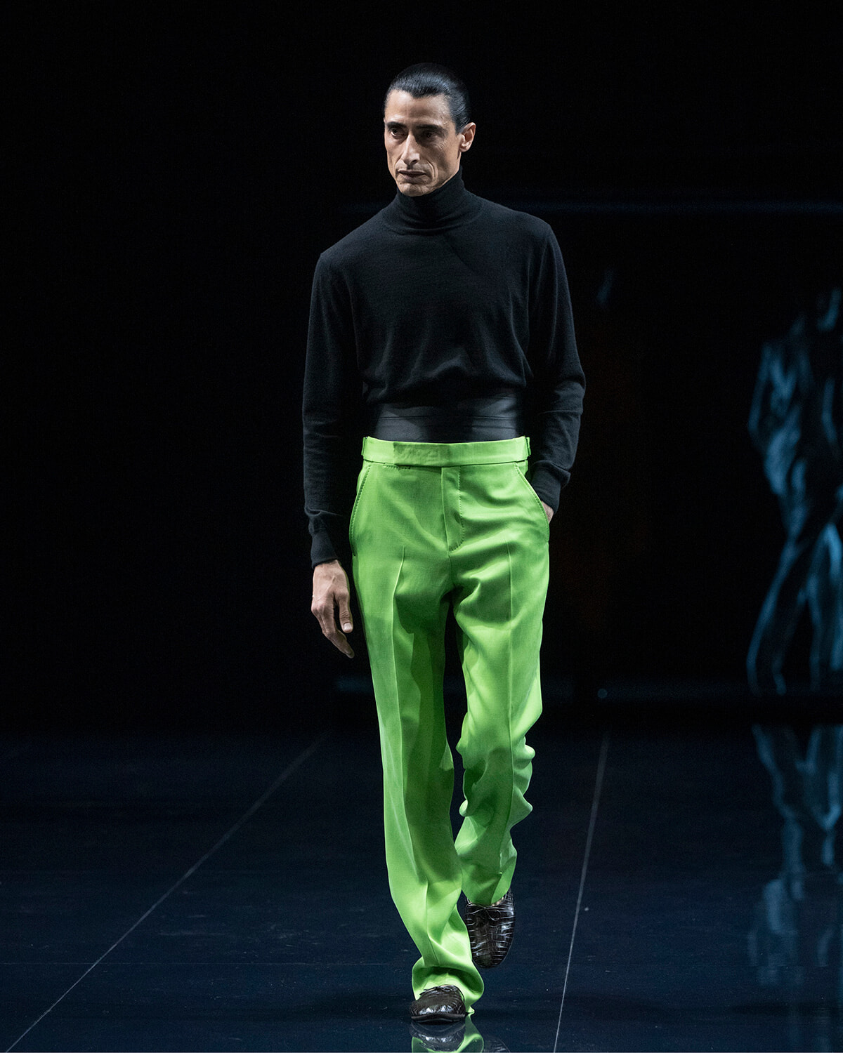 Male model walking the runway in a black turtleneck and bright lime-green trousers on a dark stage.