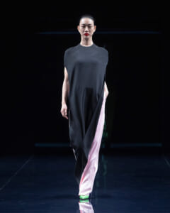 Model walking the runway in a loose black tunic top with pink wide-leg pants and green shoes on a dark stage