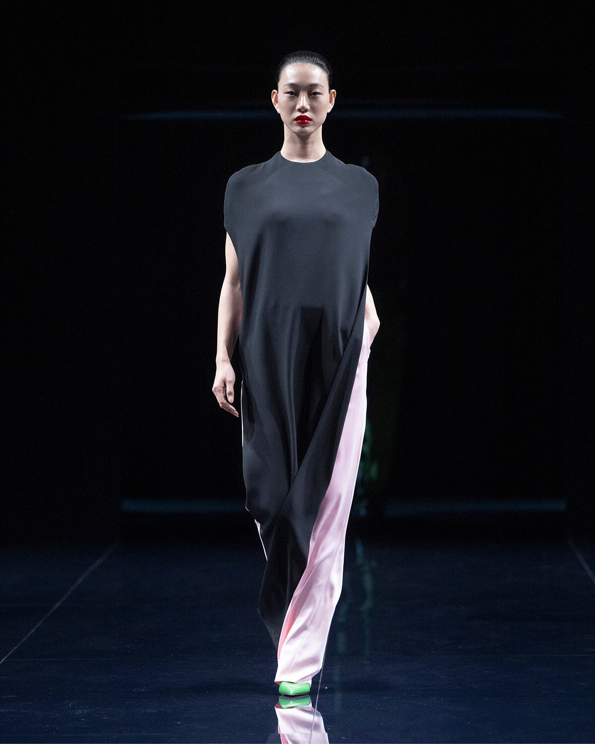 Model walking the runway in a loose black tunic top with pink wide-leg pants and green shoes on a dark stage