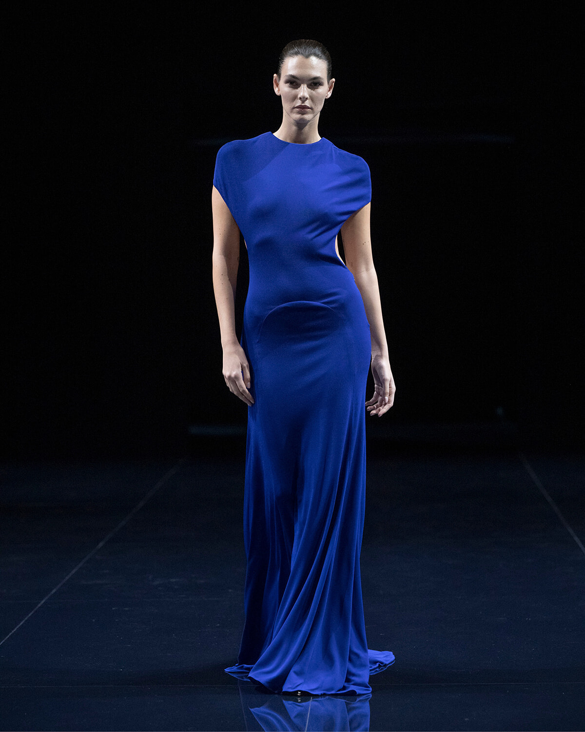 Model walking on a dark runway wearing a floor-length royal blue gown with short sleeves and a side cutout detail.