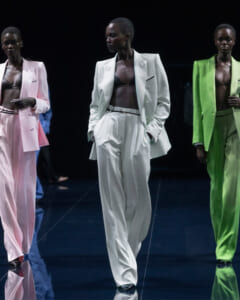 Runway models walk a fashion show wearing oversized pastel suits—white, pink, and lime green—with black bras underneath.