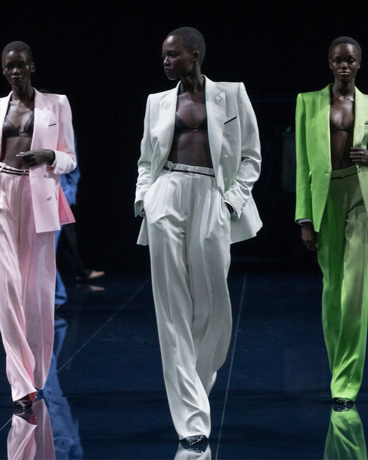 Runway models walk a fashion show wearing oversized pastel suits—white, pink, and lime green—with black bras underneath.