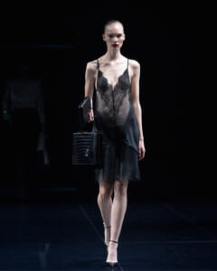 Model walks the runway in a sheer black lace slip dress, carrying a textured black handbag and a small black clutch.