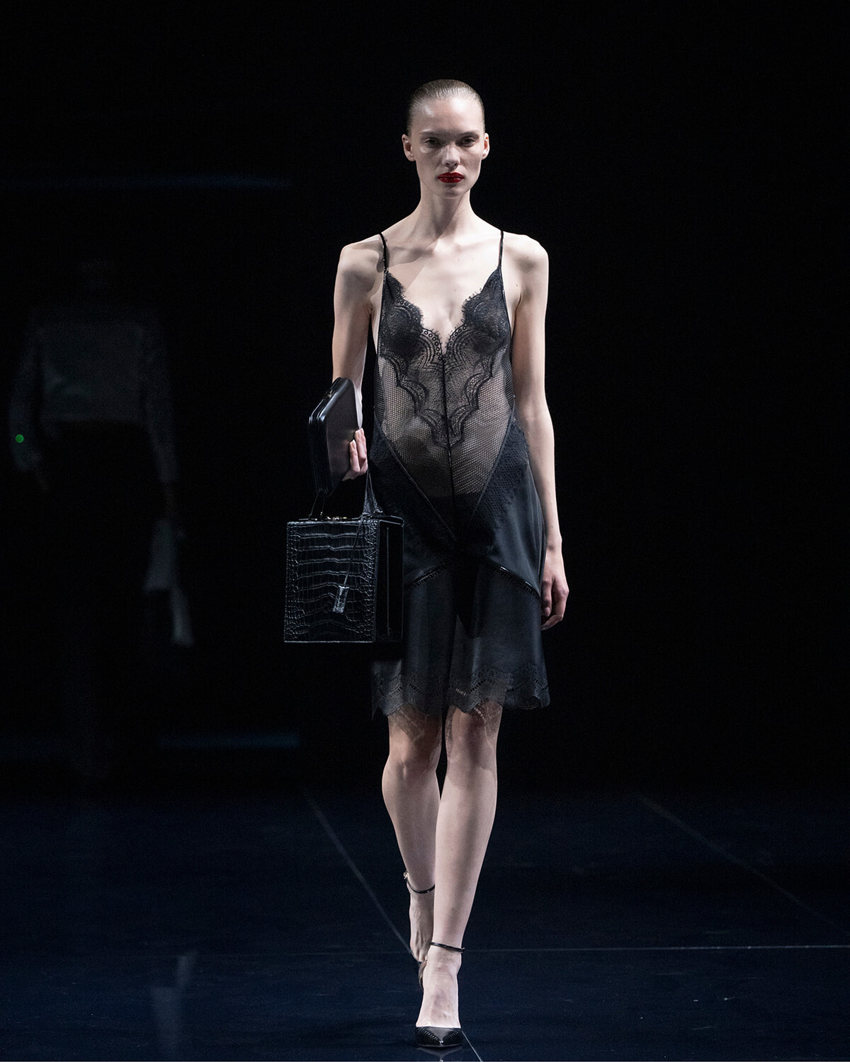 Model walks the runway in a sheer black lace slip dress, carrying a textured black handbag and a small black clutch.