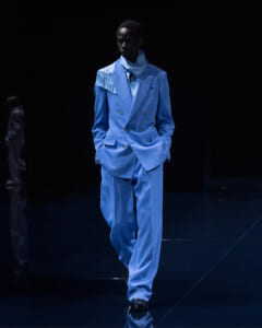 Male model walking a fashion runway in a light blue double-breasted suit with fringe detail on the left shoulder, scarf, and black shoes against a dark backdrop.