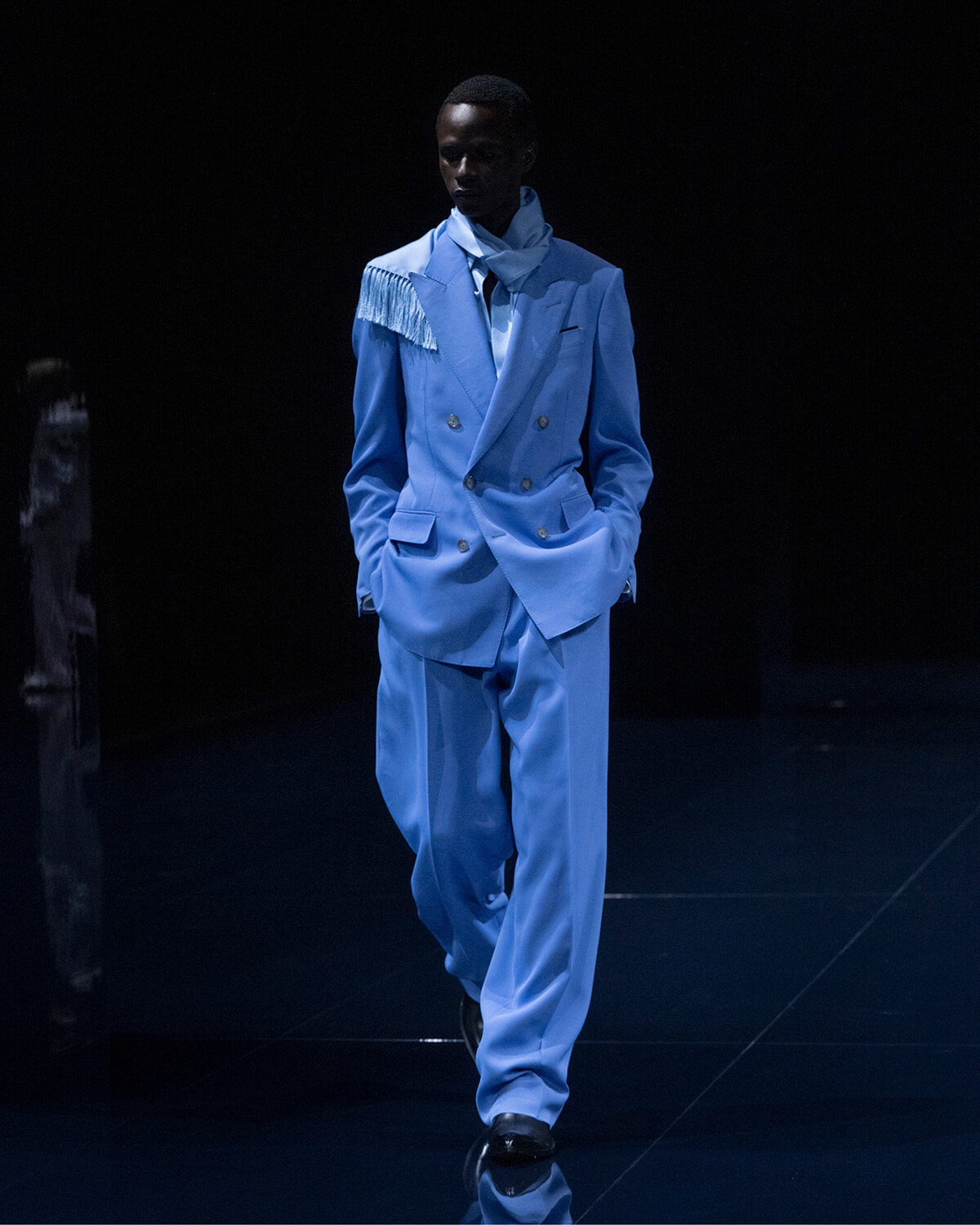 Male model walking a fashion runway in a light blue double-breasted suit with fringe detail on the left shoulder, scarf, and black shoes against a dark backdrop.