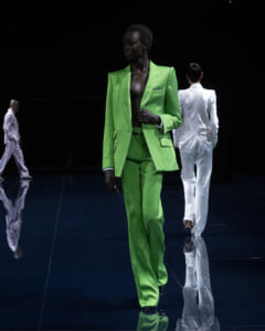 Model in a vivid lime-green suit walks the runway, with white-suited models in the background on a glossy dark floor.