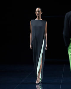 Female model walking a runway in a dark, sleeveless dress with a mint green inner panel and a thigh-high slit, on a black stage.