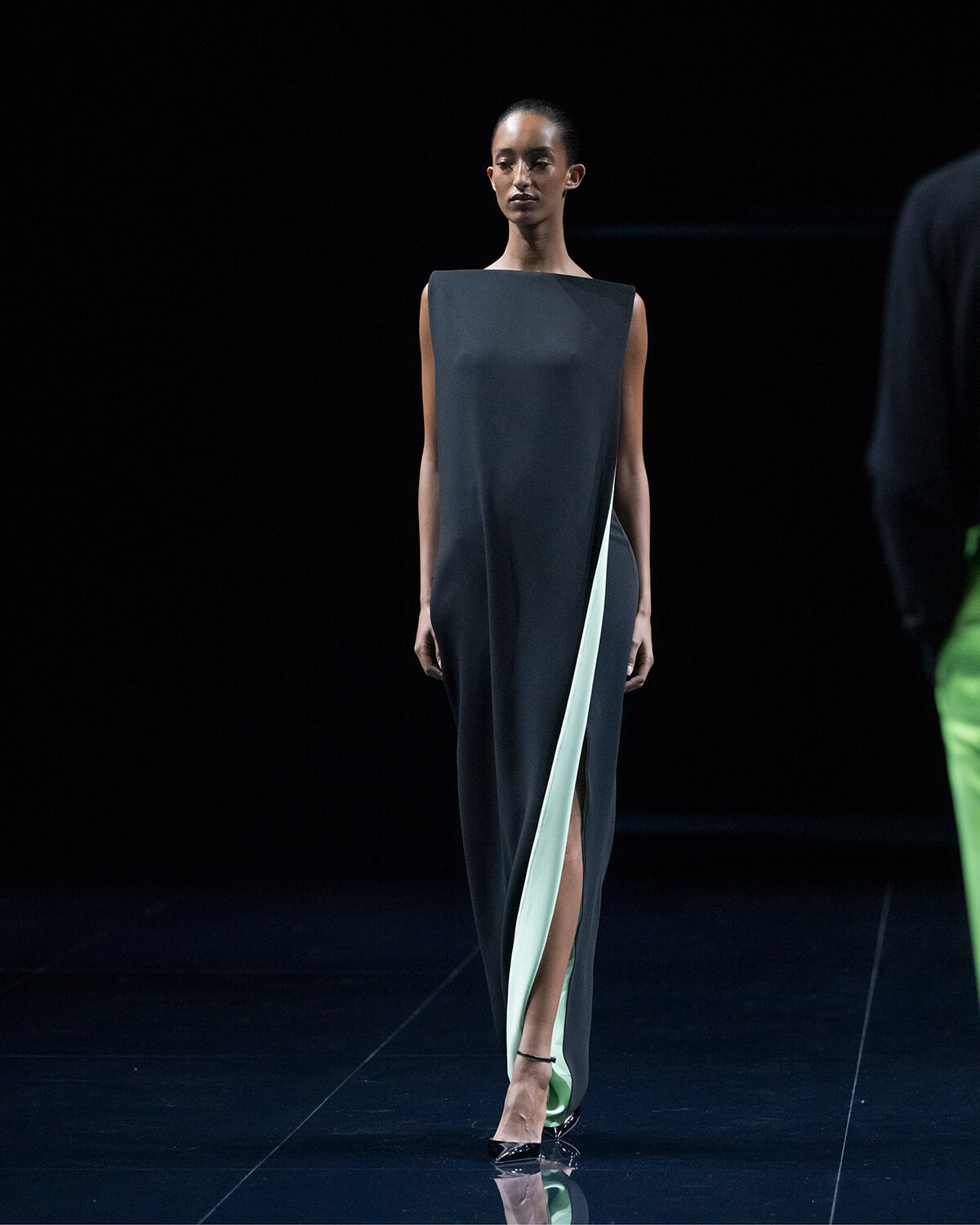 Female model walking a runway in a dark, sleeveless dress with a mint green inner panel and a thigh-high slit, on a black stage.