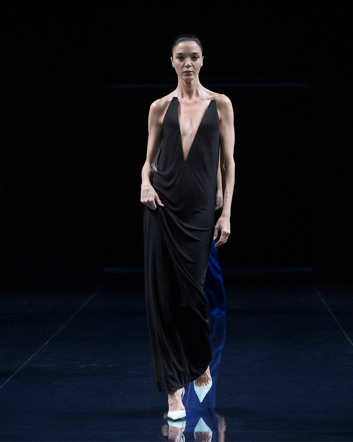 Model walking a dark runway in a black deep-V halter dress with white pointed heels.