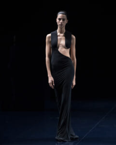 Model on a dark runway wearing a floor-length black gown with an asymmetric cutout revealing the midriff and minimalist shoulder panels.