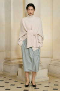 Model in a chunky cream turtleneck sweater tied at the waist with a pale blue midi skirt, standing in a stone arcade.