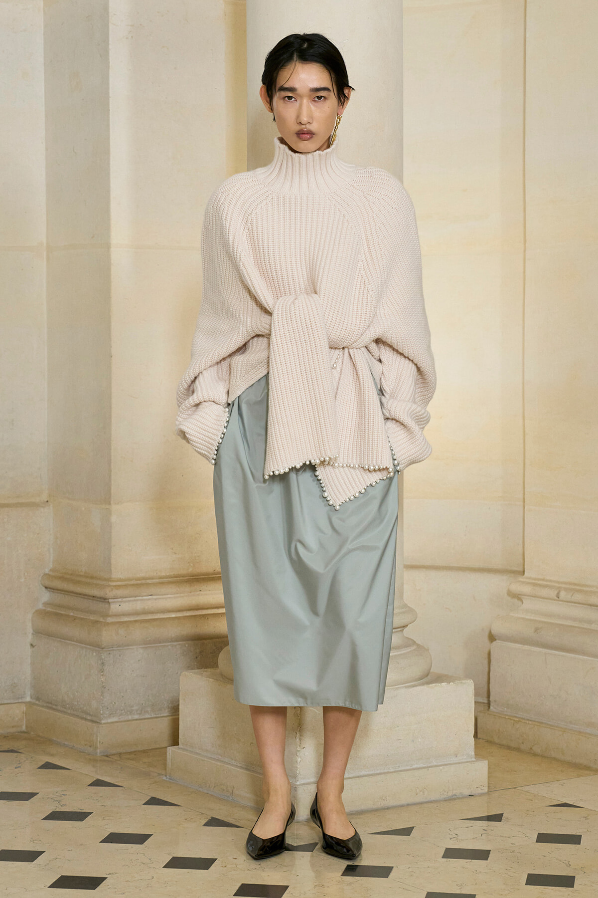 Model in a chunky cream turtleneck sweater tied at the waist with a pale blue midi skirt, standing in a stone arcade.