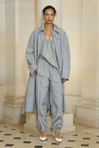 Model wearing an oversized light gray trench coat over a pale green top and matching gray pants, standing by cream architectural columns with white flats.