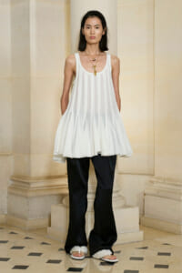 Model wearing a white pleated sleeveless tunic over black wide-leg pants, with layered gold necklaces and pearl-embellished sandals.