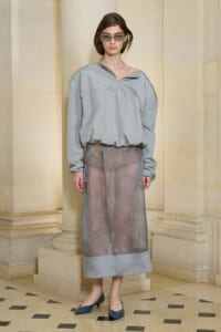 Female model in a light gray oversized top with a zipper at the neckline paired with a sheer gray midi skirt featuring pink embroidery, standing in a stone-walled setting.