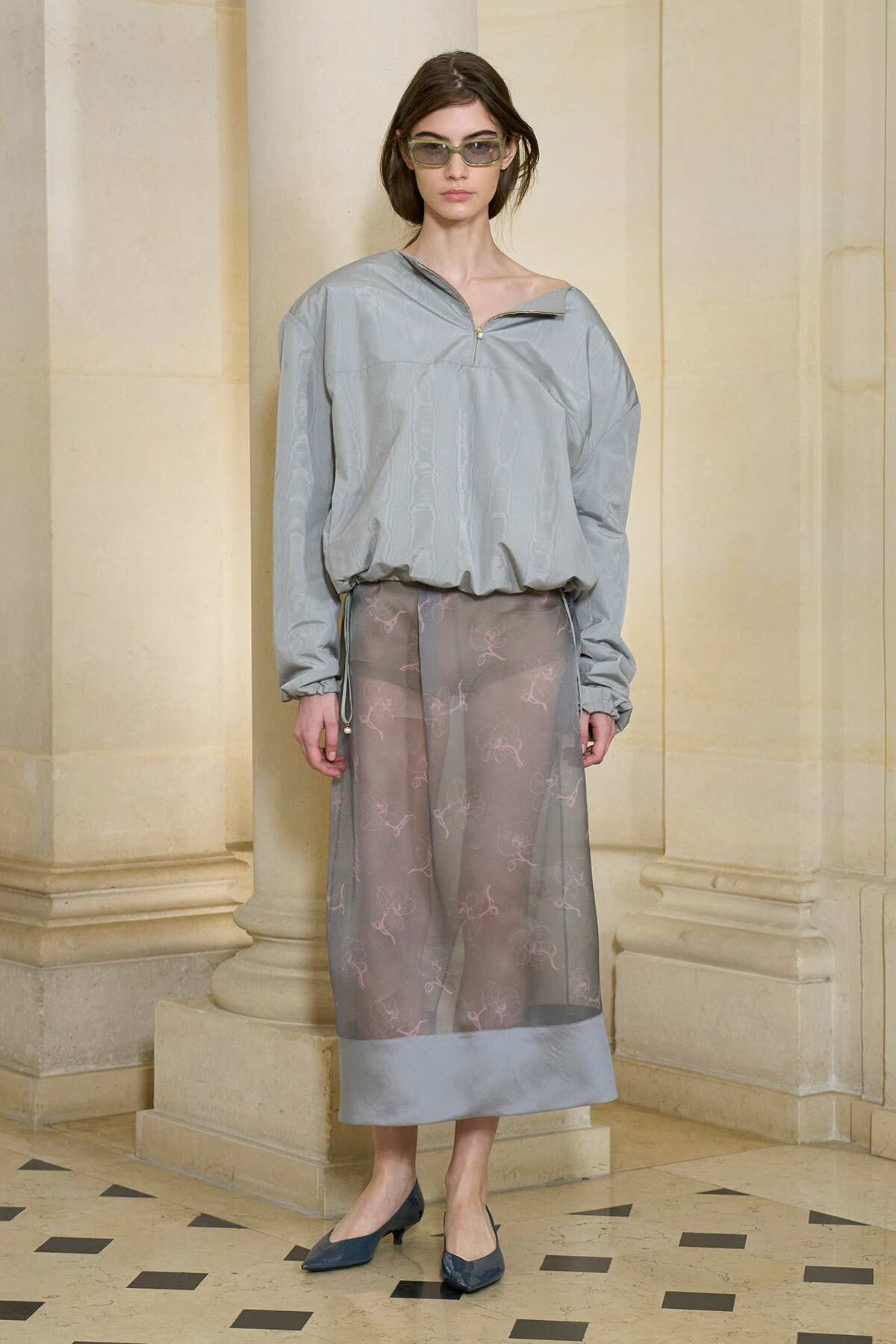 Female model in a light gray oversized top with a zipper at the neckline paired with a sheer gray midi skirt featuring pink embroidery, standing in a stone-walled setting.