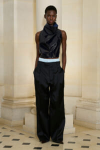 Model wearing a glossy black sleeveless high-neck crop top and oversized black trousers with a light blue waistband, standing in a stone hallway.