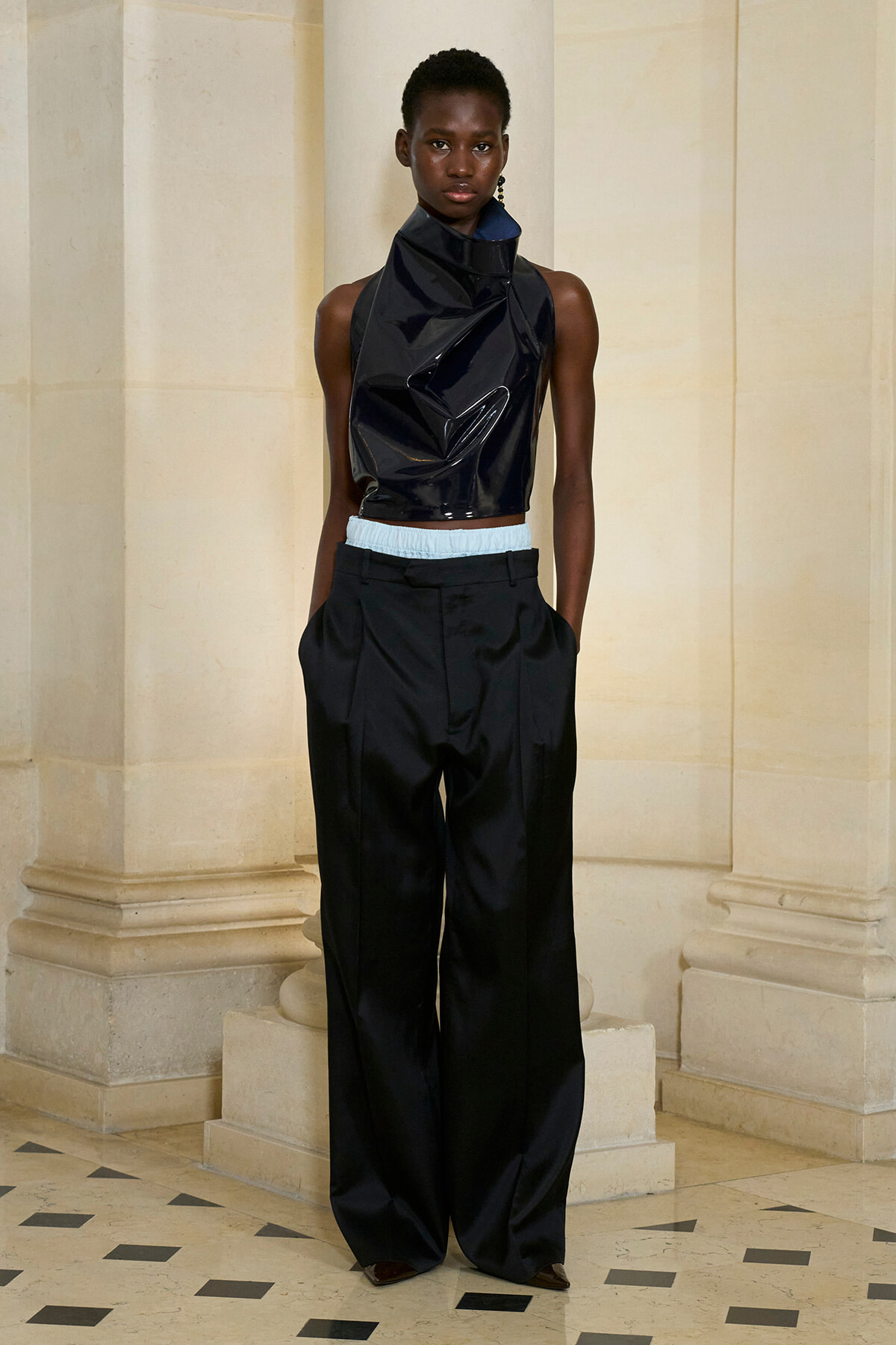 Model wearing a glossy black sleeveless high-neck crop top and oversized black trousers with a light blue waistband, standing in a stone hallway.