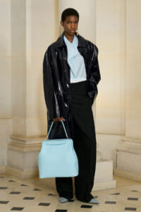 Model wearing a glossy black trench coat over a light blue top and black wide-leg pants, carrying a pale blue handbag in a columned hallway.