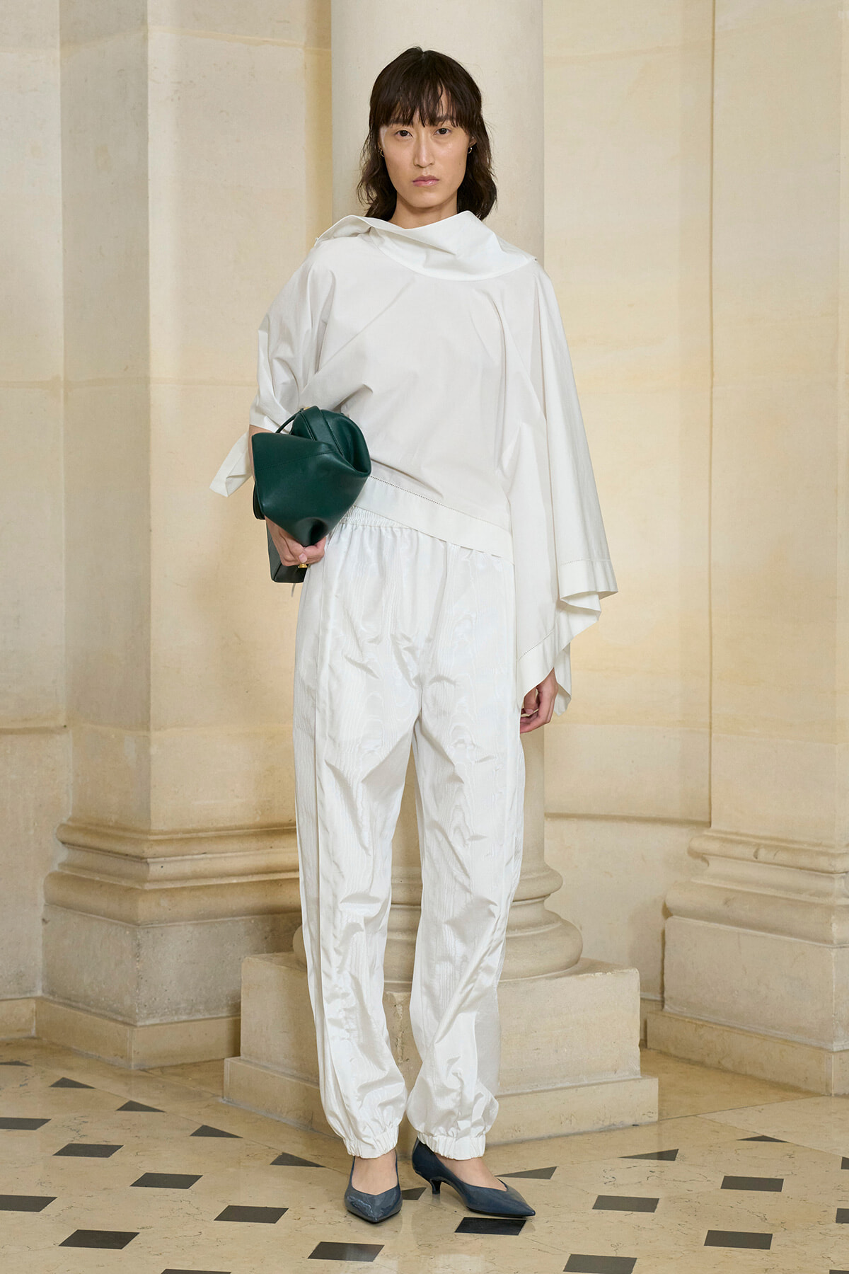 Woman in an all-white, loose-fitting outfit holding a dark green clutch, standing against beige columns indoors.