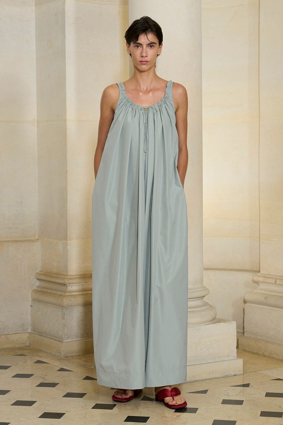Woman in a pale blue, loose halter-neck maxi dress standing between stone columns in a classical interior, hands at sides.