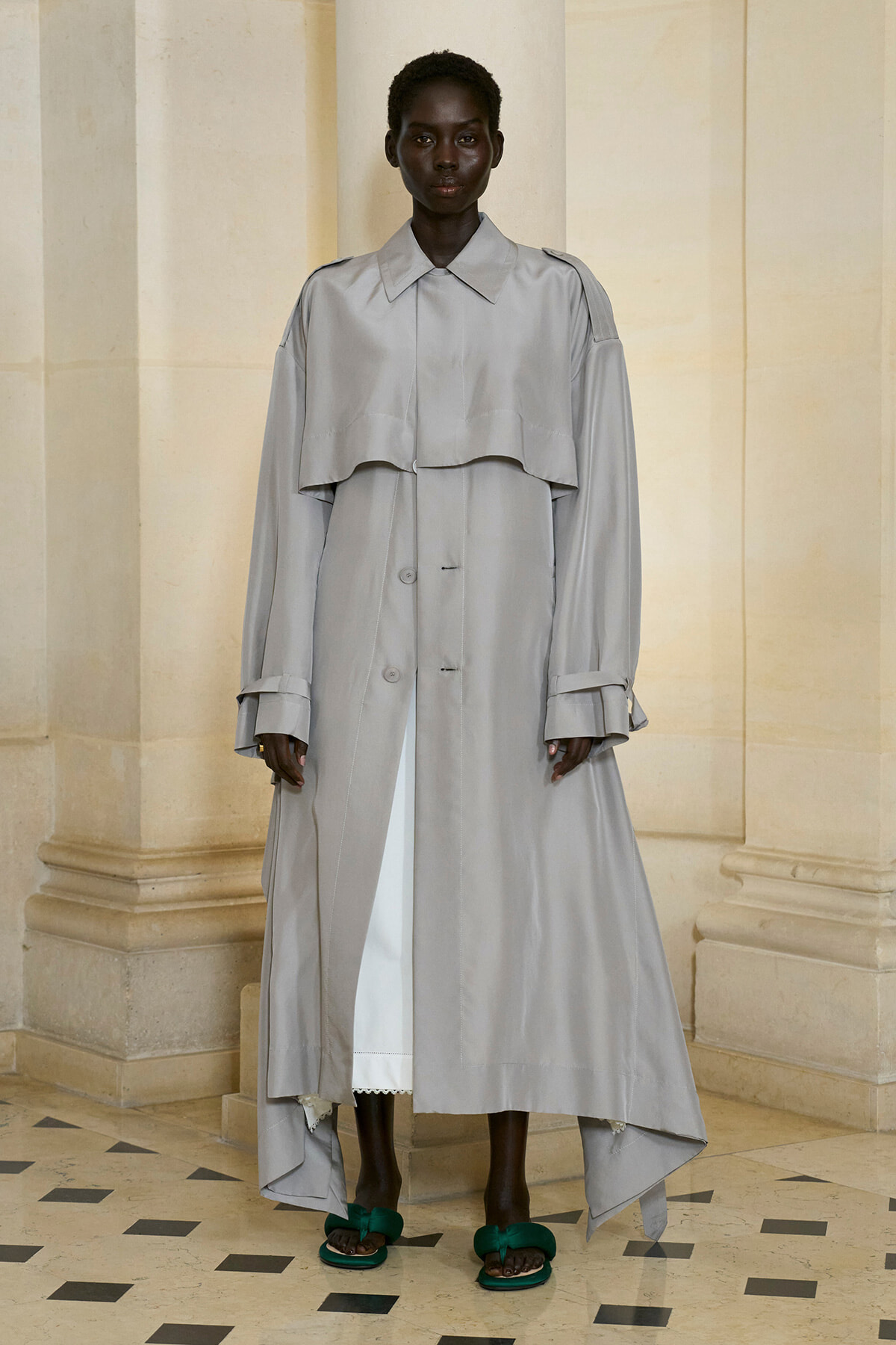 Model wearing an oversized light grey trench coat with a cape-like yoke, standing indoors against columns; dark skin and short hair, no belt, green sandals visible.