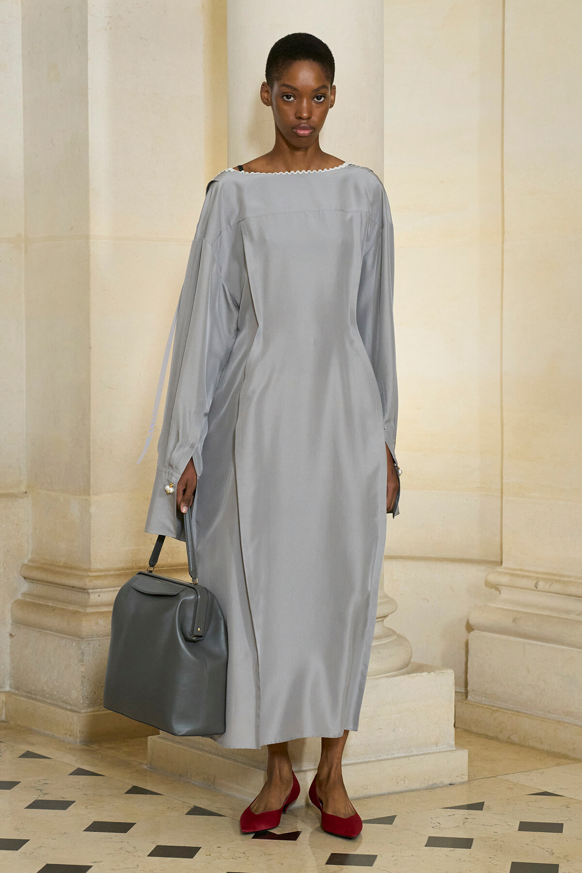 Model in a pale gray, long-sleeve, ankle-length dress with a boat neckline, carrying a gray handbag and wearing red flats, standing beside columned architecture.