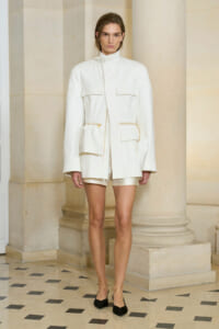 Model wearing an oversized white utility jacket with large front pockets, standing in a marble hallway.