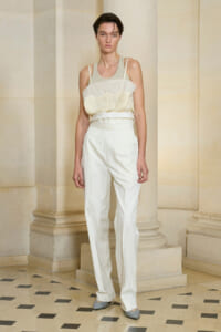 Model wearing a cream lace-trimmed camisole tucked into high-waisted white trousers, standing against columned architecture.