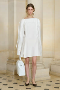 Model wearing a loose white shift dress, standing by a stone column, holding a white bag and wearing brown flats in a beige architectural setting.